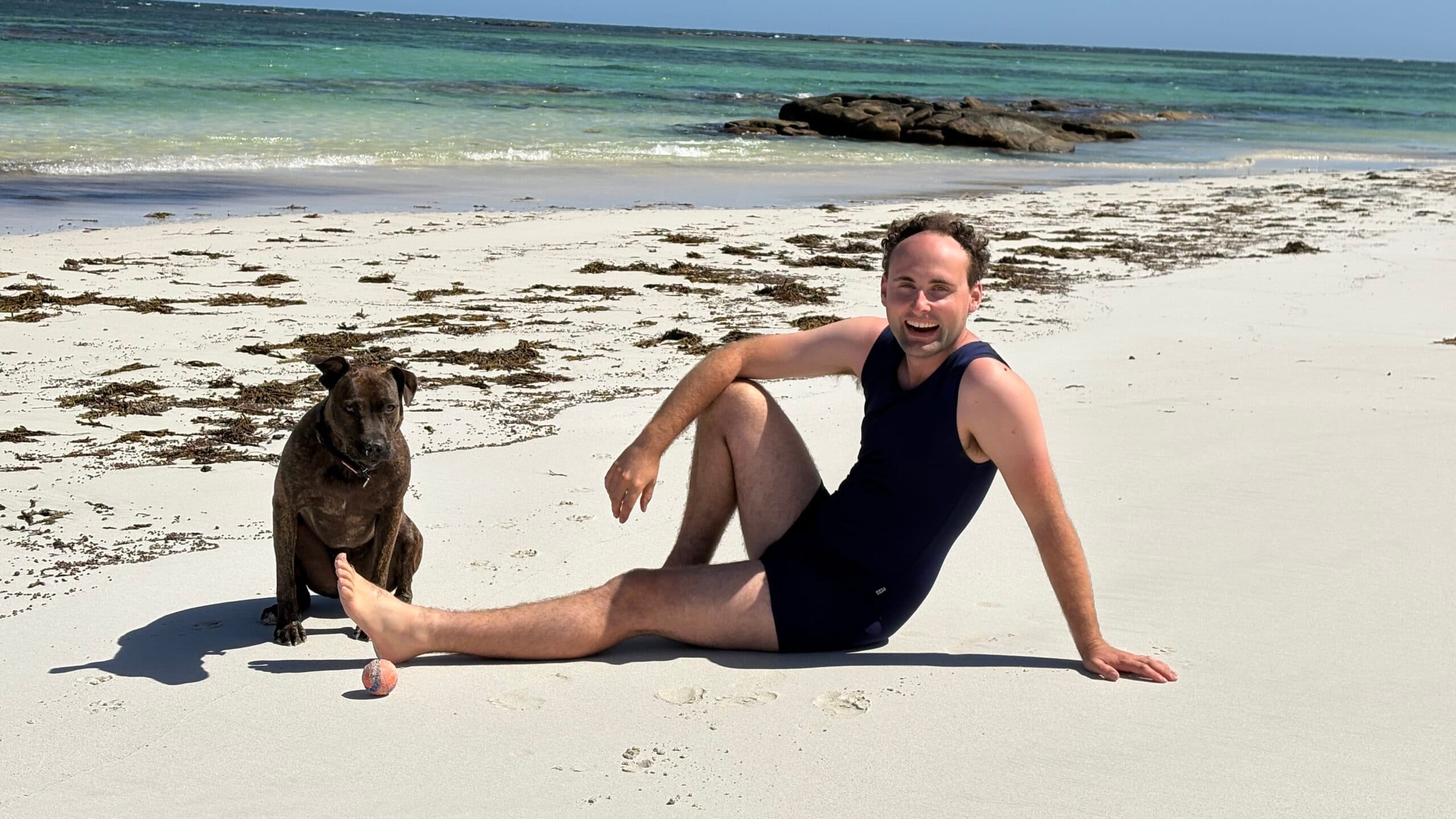 Model wearing a black one-piece swimsuit beside a dog on the beach